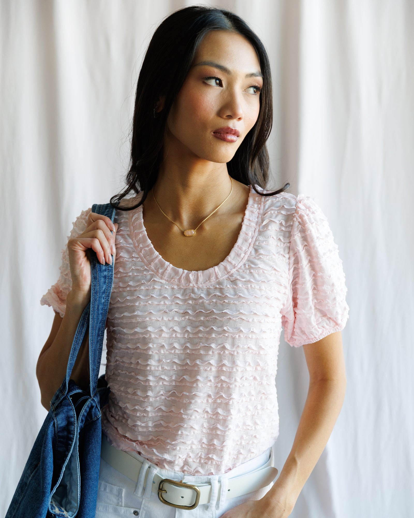 Woman wearing a light pink textured top holding a denim bag against a white curtain background