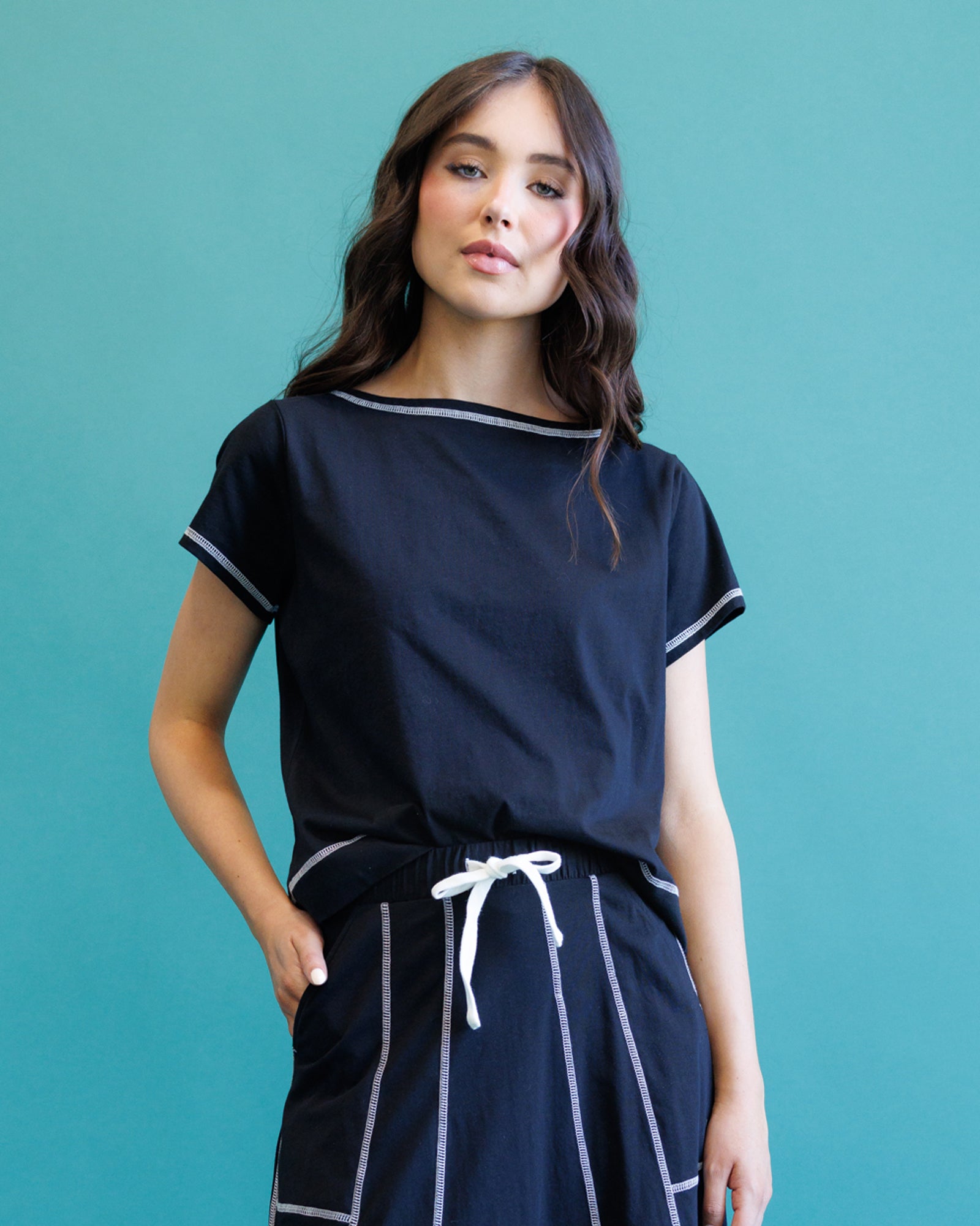 A woman modeling a navy blue short-sleeve top with white trim and matching navy skirt with white vertical stripes and a white drawstring, set against a solid teal background.