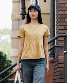 A woman wearing a mustard yellow short-sleeve peplum top, blue jeans, and a navy blue New York Yankees baseball cap, standing on outdoor stairs.