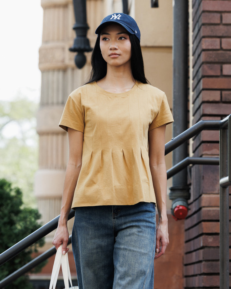 A woman wearing a mustard yellow short-sleeve peplum top, blue jeans, and a navy blue New York Yankees baseball cap, standing on outdoor stairs.