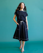 A woman modeling a navy blue two-piece set consisting of a short-sleeve top and a matching midi skirt with white contrast stitching and a white drawstring, set against a solid teal background.