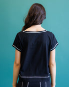 A woman viewed from behind wearing a short-sleeve black top with thin white contrast trim along the neckline, sleeve cuffs, and hem, against a solid teal background.