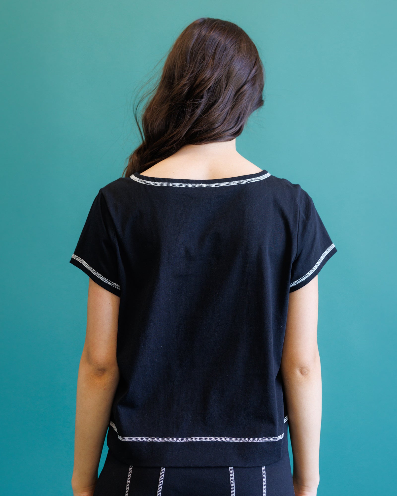 A woman viewed from behind wearing a short-sleeve black top with thin white contrast trim along the neckline, sleeve cuffs, and hem, against a solid teal background.