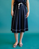 A model wearing a black midi skirt with white vertical stripes and a white drawstring waist, paired with flat shoes, set against a solid teal background.