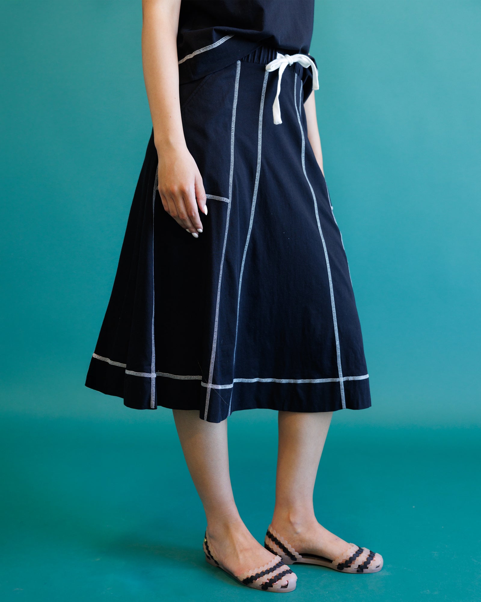 A person wearing a dark navy midi skirt with white contrast stitching and a white drawstring waist, paired with black and white flat sandals, set against a solid teal background.
