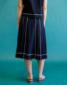 A person shown from the back wearing a dark navy midi skirt with white vertical and horizontal stripe details, paired with a matching dark top, against a solid teal background.