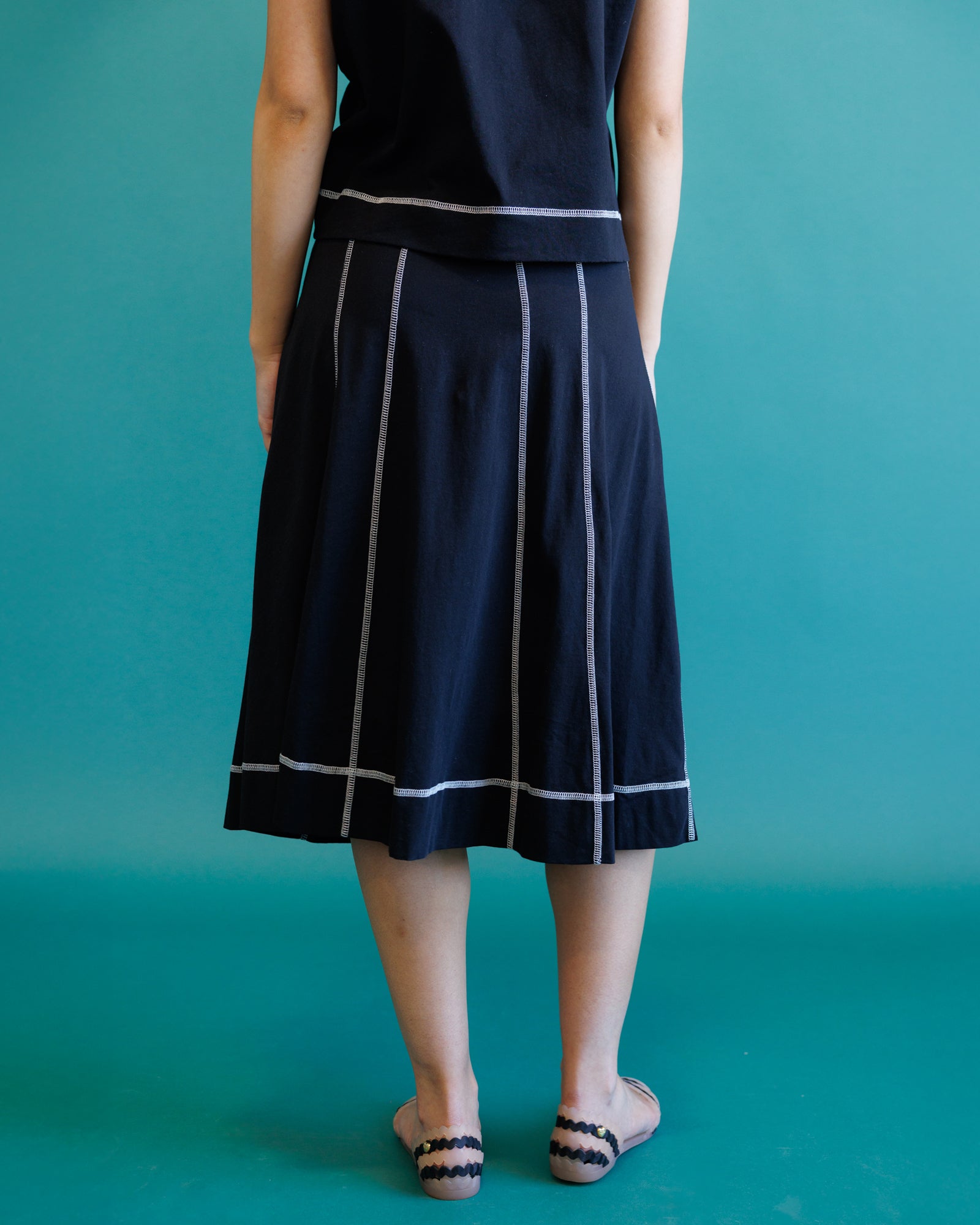 A person shown from the back wearing a dark navy midi skirt with white vertical and horizontal stripe details, paired with a matching dark top, against a solid teal background.