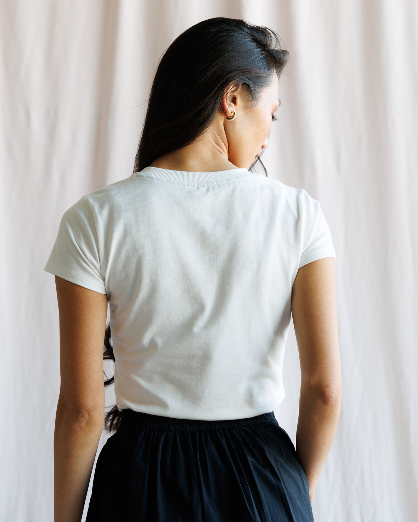 Person wearing a white t-shirt and dark skirt against a light curtain background