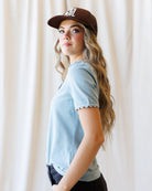 A woman wearing a light blue short-sleeve t-shirt with scalloped edges and a brown baseball cap, photographed against a soft cream-colored curtain backdrop.