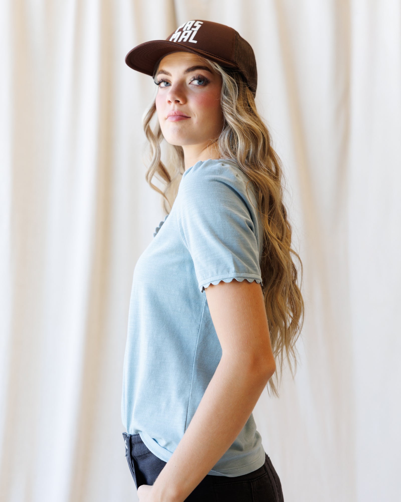 A woman wearing a light blue short-sleeve t-shirt with scalloped edges and a brown baseball cap, photographed against a soft cream-colored curtain backdrop.