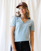A woman wearing a light blue short-sleeve V-neck t-shirt with scalloped edges, paired with dark jeans and a brown baseball cap with white text, standing against a soft white curtain background.