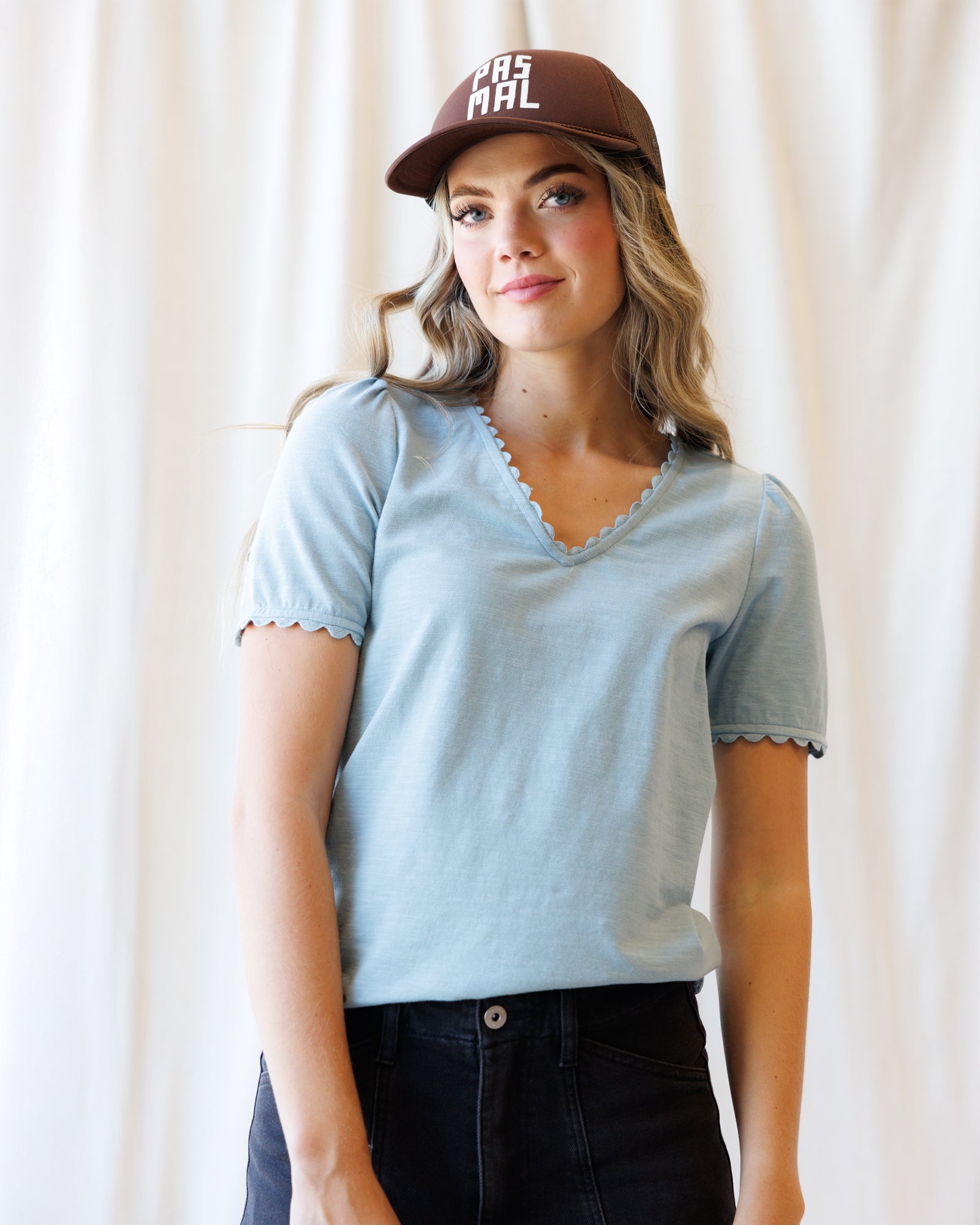 A woman wearing a light blue short-sleeve V-neck t-shirt with scalloped edges, paired with dark jeans and a brown baseball cap with white text, standing against a soft white curtain background.