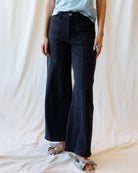 A person wearing black wide-leg denim pants with a light blue top and patterned flat sandals, standing against a draped cream fabric backdrop.