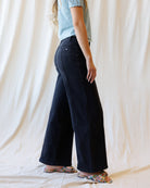 A person shown from the waist down wearing dark wide-leg denim pants, a light blue t-shirt, and colorful plaid slide sandals, standing against a draped cream fabric backdrop.