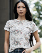 A woman wearing a short-sleeve crewneck t-shirt with a faded vintage floral pattern in muted pink, blue, and green tones on a light beige background, paired with dark blue jeans.