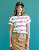 A woman modeling a white short-sleeve crewneck t-shirt with thin horizontal stripes in green, pink, and blue, paired with a brown baseball cap and a tan skirt, set against a solid teal background.