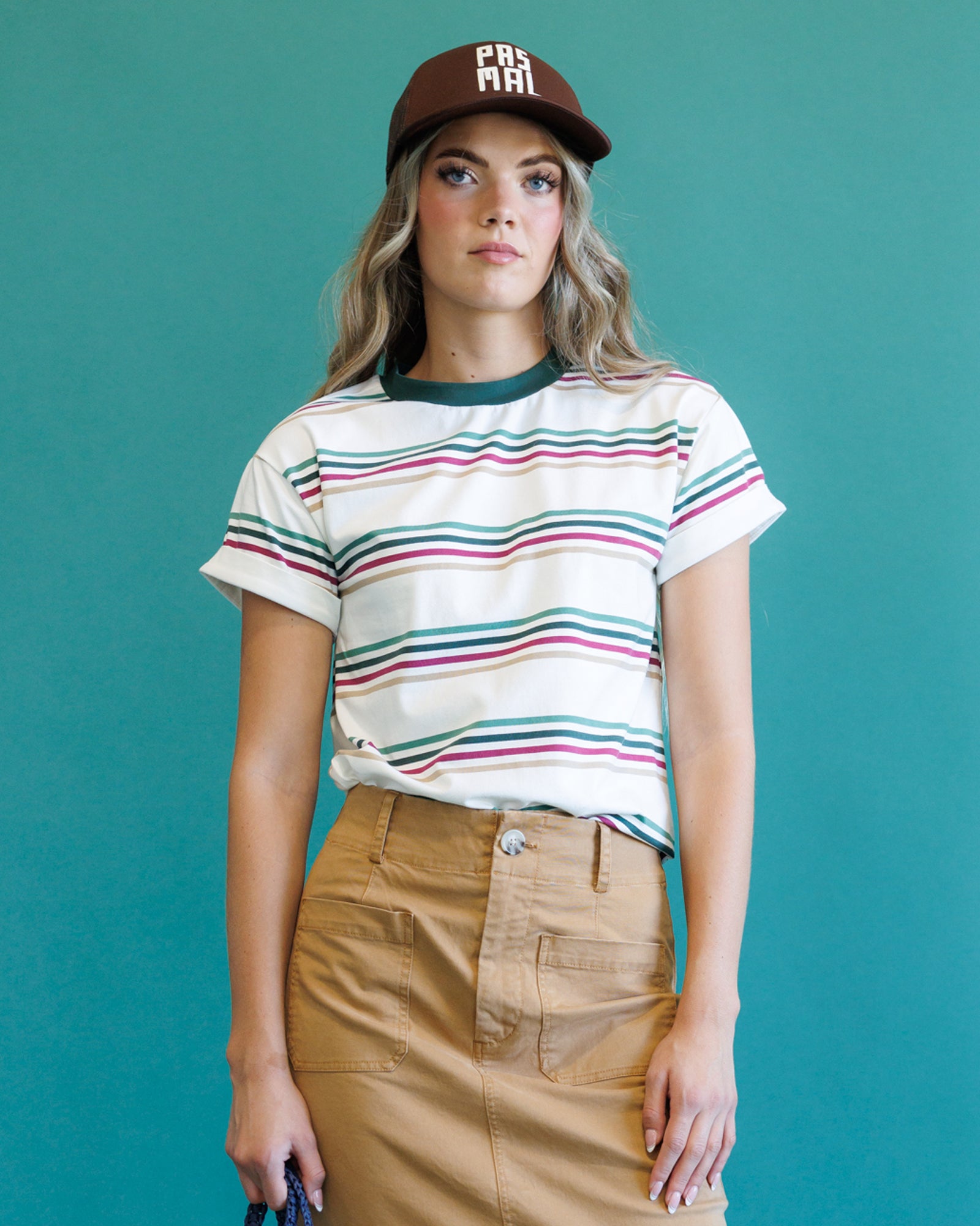 A woman modeling a white short-sleeve crewneck t-shirt with thin horizontal stripes in green, pink, and blue, paired with a brown baseball cap and a tan skirt, set against a solid teal background.