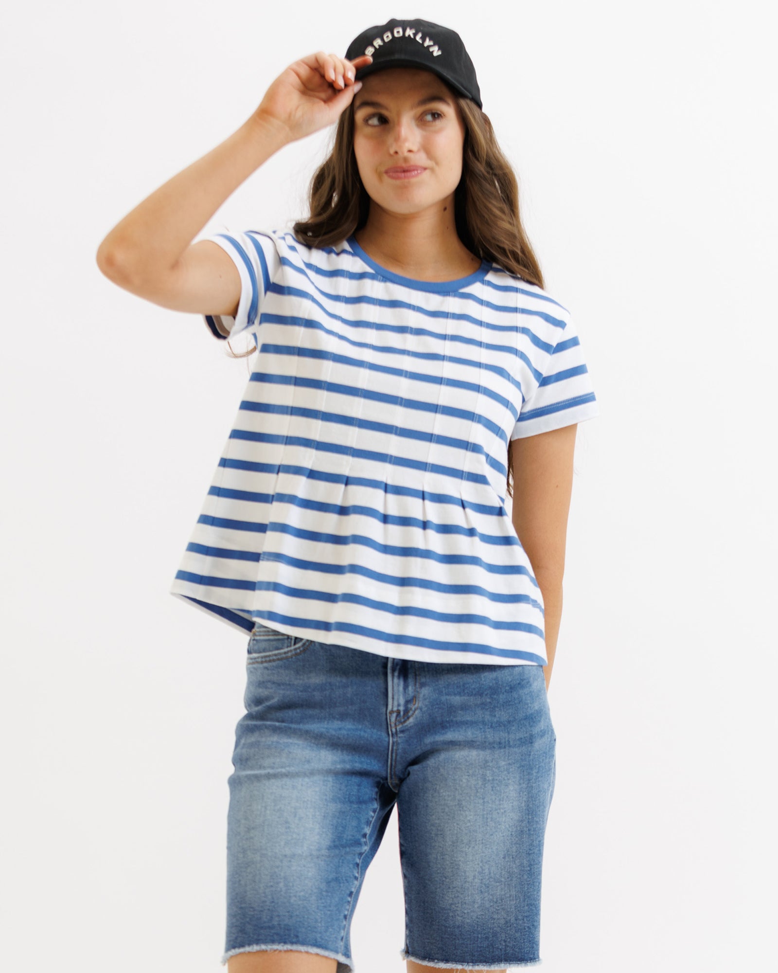 Woman wearing a blue and white striped shirt, denim shorts, and a black cap with text on a white background