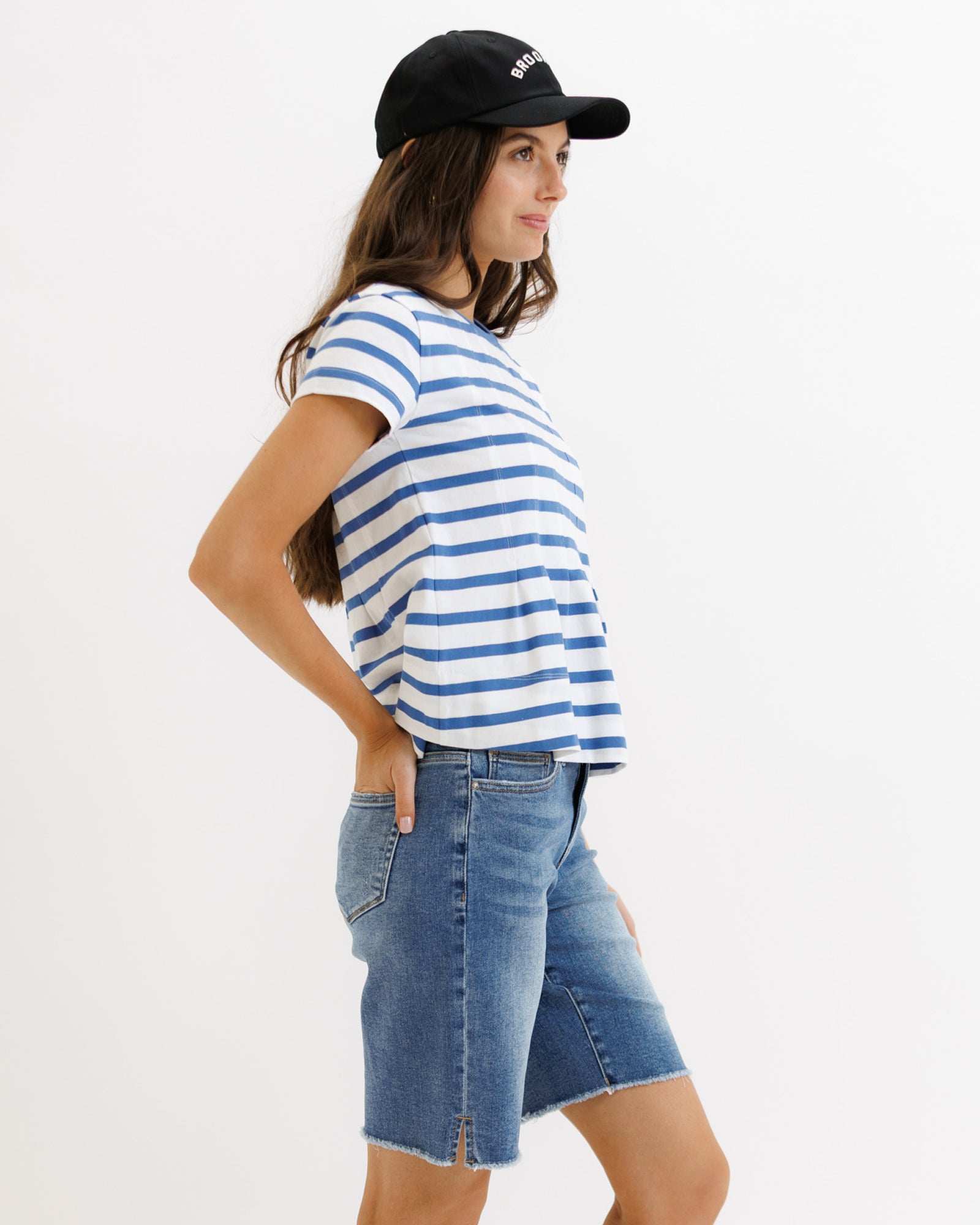 Woman wearing a blue and white striped shirt, denim shorts, and a black cap on a white background