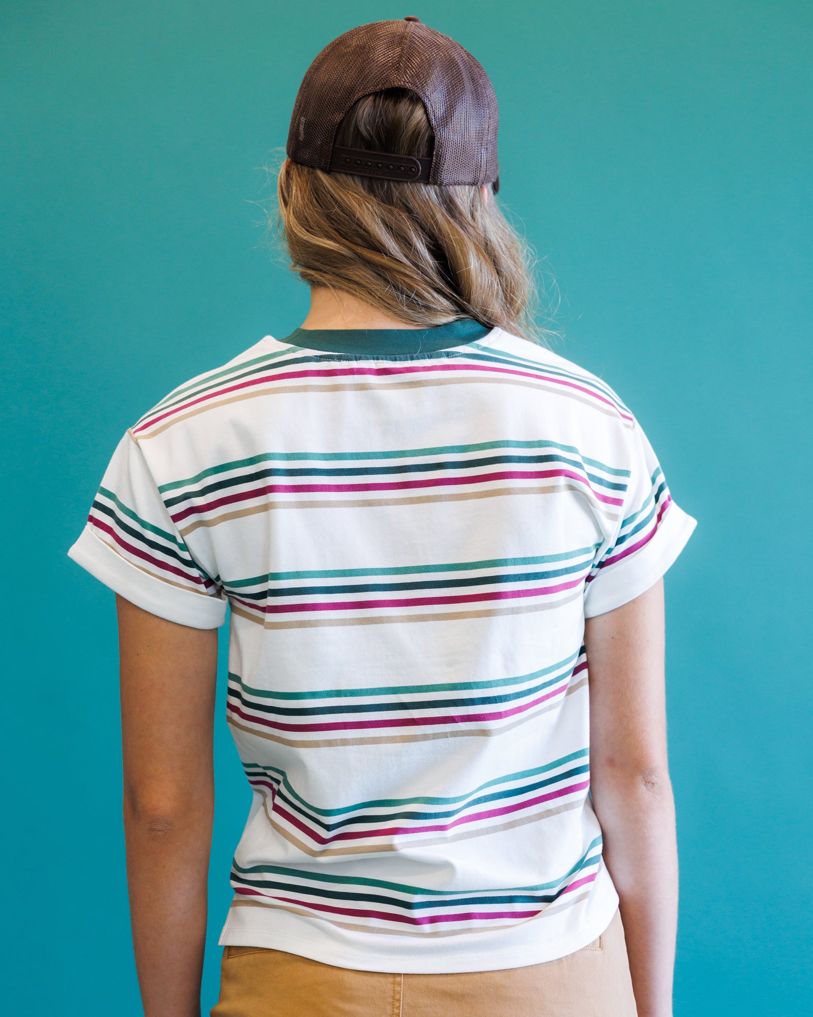 A person viewed from behind wearing a white short-sleeve ringer T-shirt with thin horizontal stripes in teal, pink, and tan, paired with a brown baseball cap and tan pants, set against a solid teal background.