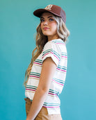 A woman wearing a white short-sleeve t-shirt with thin pink, green, and blue horizontal stripes, paired with a brown baseball cap and tan pants, set against a solid teal background.