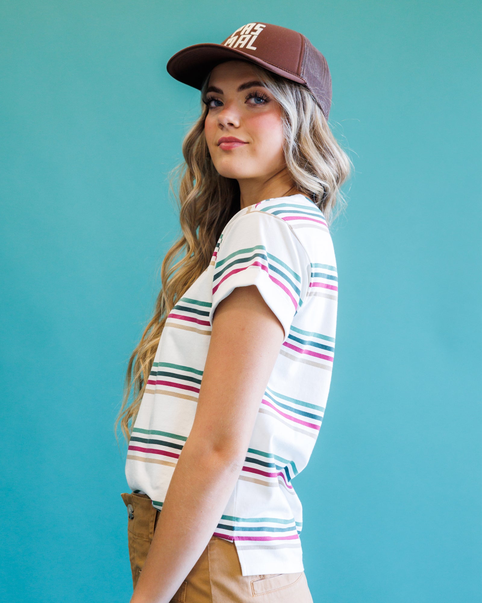 A woman wearing a white short-sleeve t-shirt with thin pink, green, and blue horizontal stripes, paired with a brown baseball cap and tan pants, set against a solid teal background.