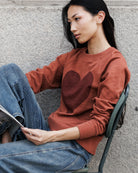 A woman sitting on a metal chair against a gray concrete wall, wearing a rust-orange crewneck sweatshirt with a dark brown heart graphic on the chest and blue jeans.
