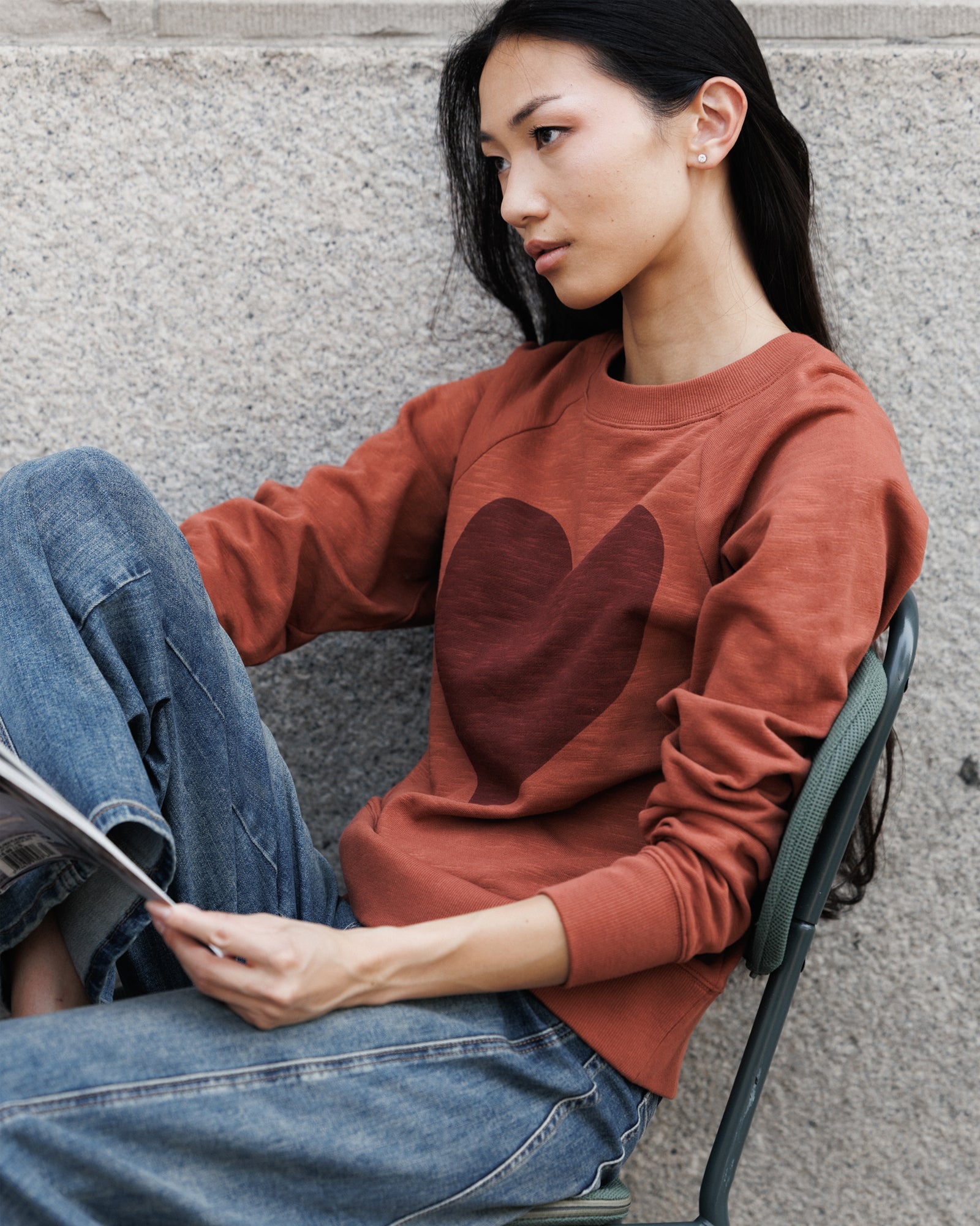 A woman sitting on a metal chair against a gray concrete wall, wearing a rust-orange crewneck sweatshirt with a dark brown heart graphic on the chest and blue jeans.