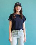 A woman wearing a navy blue short-sleeve ringer T-shirt with white trim, light-wash jeans, and a black baseball cap with 'BROOKLYN' in white text, standing against a solid teal background.