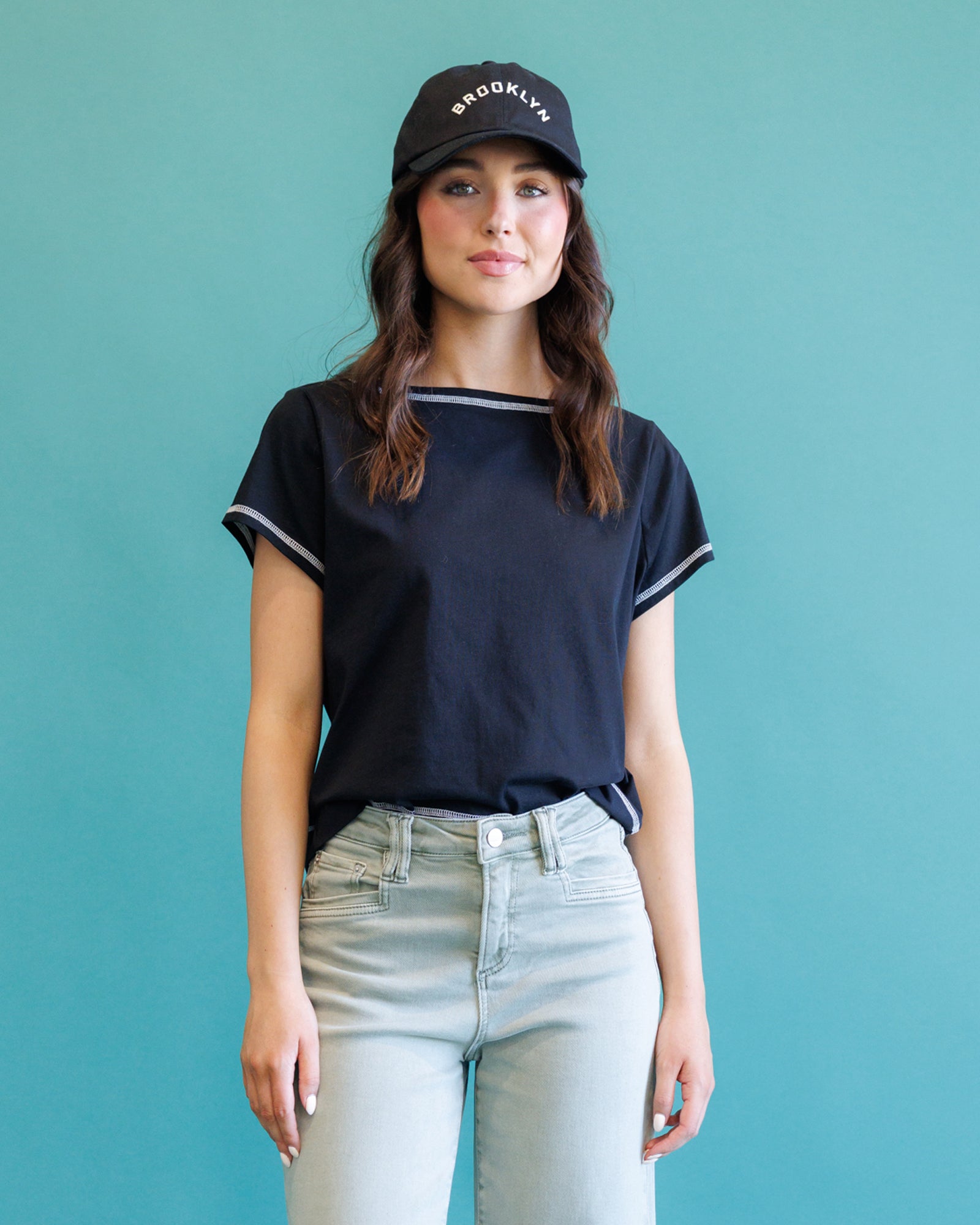 A woman wearing a navy blue short-sleeve ringer T-shirt with white trim, light-wash jeans, and a black baseball cap with 'BROOKLYN' in white text, standing against a solid teal background.