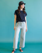 A woman modeling a navy blue ringer t-shirt with white trim, light-wash wide-leg jeans, a black baseball cap, and dark brown slide sandals, set against a solid teal background.
