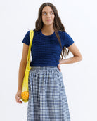 Woman wearing a blue textured top and checkered skirt, holding a yellow bag with groceries.