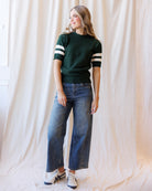 A woman modeling a dark green short-sleeve knit sweater with white stripes on the sleeves, paired with wide-leg blue jeans and white sneakers, standing against a draped cream fabric backdrop.