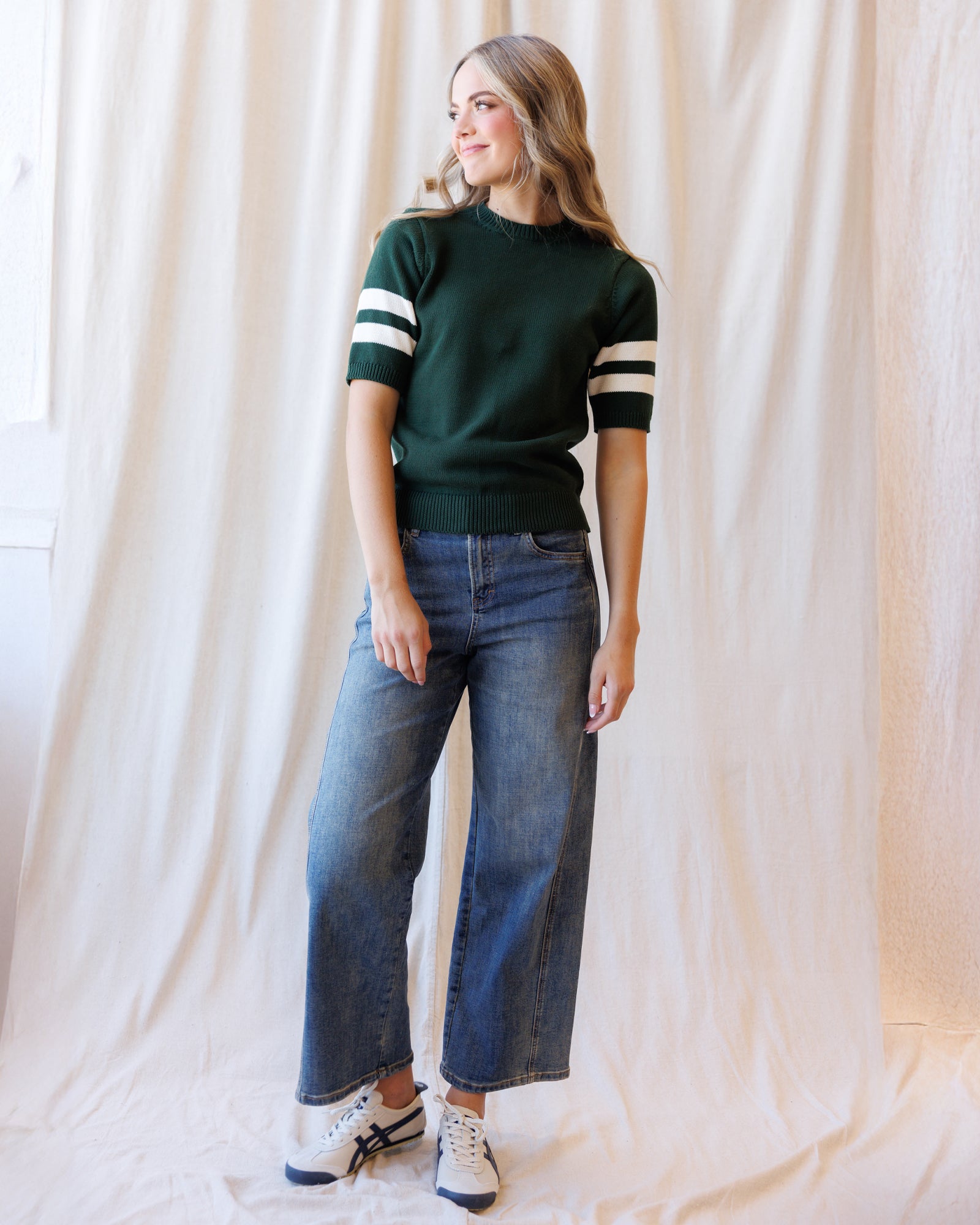 A woman modeling a dark green short-sleeve knit sweater with white stripes on the sleeves, paired with wide-leg blue jeans and white sneakers, standing against a draped cream fabric backdrop.