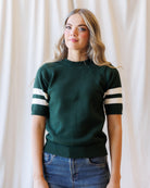 A woman wearing a dark green short-sleeve knit sweater with white stripes on the sleeves, paired with blue jeans, standing against a draped cream fabric backdrop.