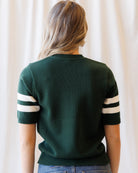 A woman viewed from behind wearing a dark green short-sleeve knit sweater with white stripes on the sleeves, paired with blue jeans against a soft cream curtain background.