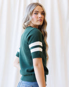 A woman wearing a dark green short-sleeve knit sweater with two white stripes on the sleeves, paired with blue jeans, standing against a soft white curtain background.