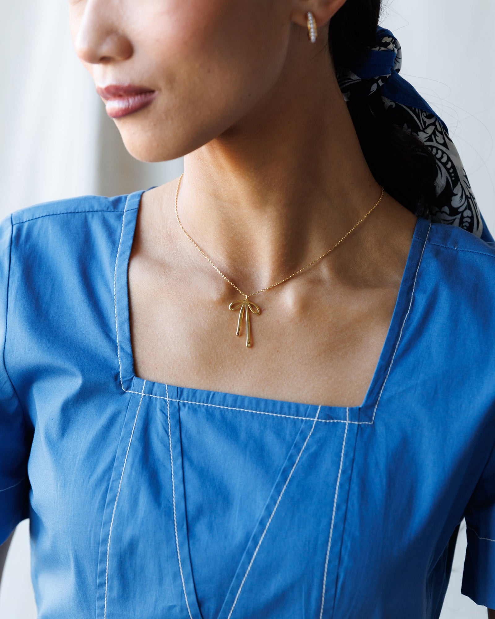 Woman wearing a gold necklace with a bow pendant, close-up on neck and blue top.
