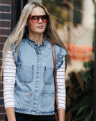 A woman wearing a light blue denim vest over a white and navy striped long-sleeve shirt, accessorized with red-framed sunglasses and a brown shoulder bag, standing outdoors.