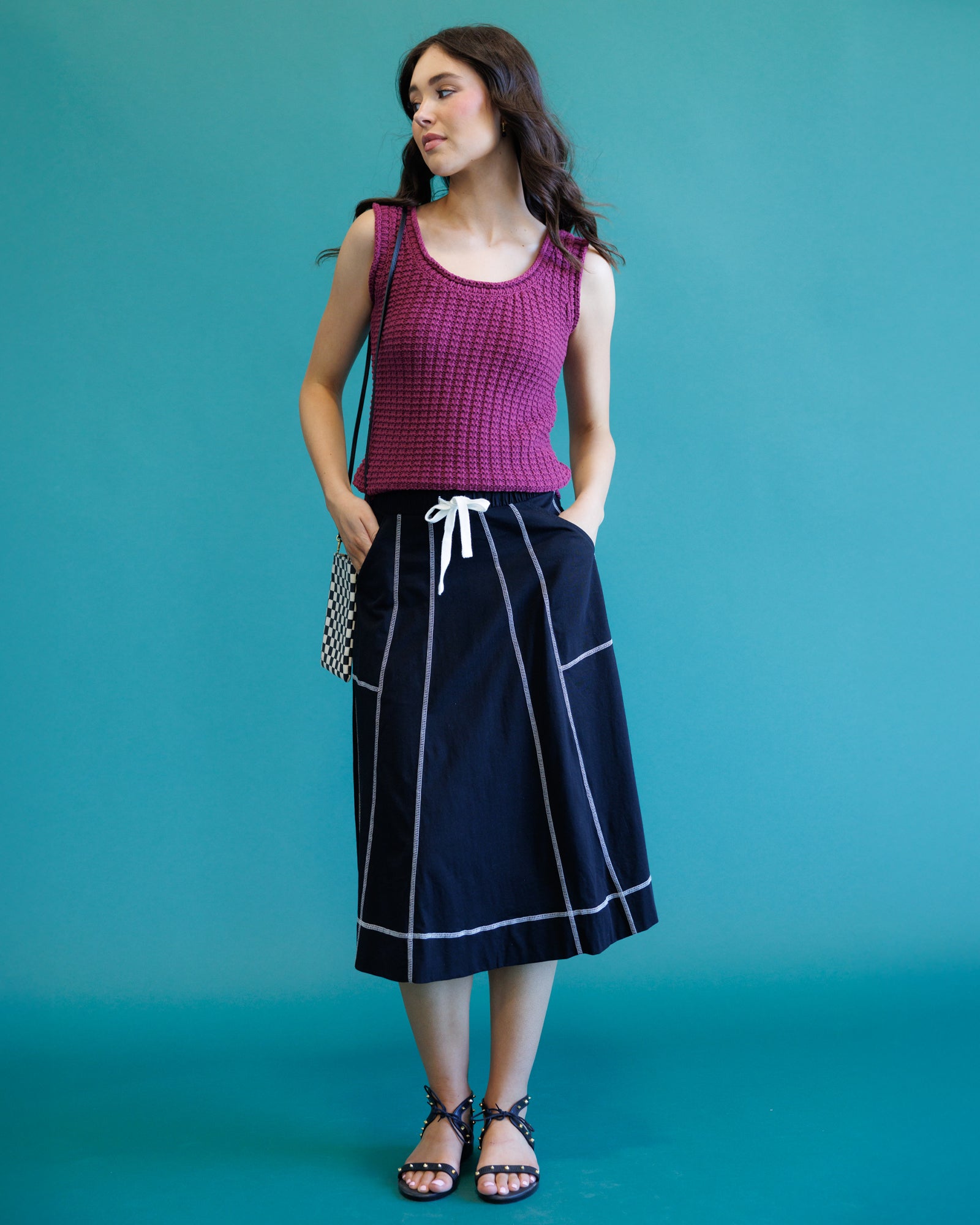A woman modeling a sleeveless magenta knit top and a dark navy midi skirt with white piping and a white drawstring, standing against a solid teal background.