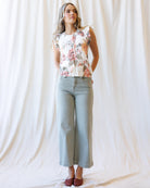 A woman stands against a draped white fabric backdrop wearing a sleeveless floral top with a cream base and pink, peach, and green floral print, paired with light blue cropped jeans and burgundy flats.