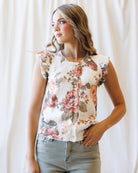 A woman wearing a sleeveless button-front top with a floral print in pink, red, and green on a cream background, paired with light gray jeans, standing against a white curtain backdrop.