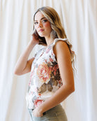A woman wearing a sleeveless white top with a large pink and brown floral print, paired with olive green pants, standing against a white curtain background.