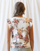 A woman viewed from behind wearing a sleeveless top with a vintage floral print in muted red, pink, and yellow tones on a cream base, paired with light gray pants against a soft white curtain background.