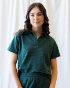 A woman modeling a dark green short-sleeve collared top with subtle horizontal stripes, paired with matching green bottoms, set against a soft white curtain background.