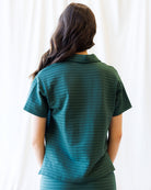 A woman viewed from behind wearing a short-sleeve collared top with a subtle horizontal striped pattern in dark teal and green, set against a soft white curtain background.