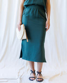 A model wearing a dark teal green striped midi skirt with a side slit, holding a woven cream clutch, and wearing black strappy sandals against a draped white fabric backdrop.