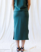A model wearing a dark teal green striped midi skirt with a side slit, and wearing black strappy sandals against a draped white fabric backdrop.