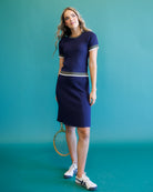A woman modeling a navy blue textured knit tennis dress with white trim on the sleeves and hem, holding a tennis racket against a solid teal background.