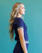 A woman in profile wearing a navy blue textured knit short-sleeve top with gray and white striped accents on the sleeve and waist, set against a solid teal background.